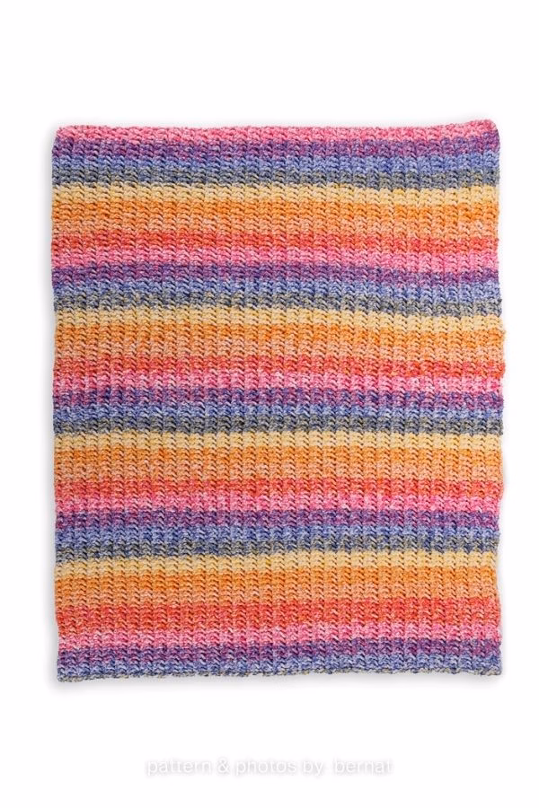 A rectangular knitted blanket laid flat against a white background, featuring horizontal stripes in a vibrant, variegated yarn with shades of pink, purple, orange, yellow, and blue, featured in a knitting roundup from https://www.theknitcrew.com/.