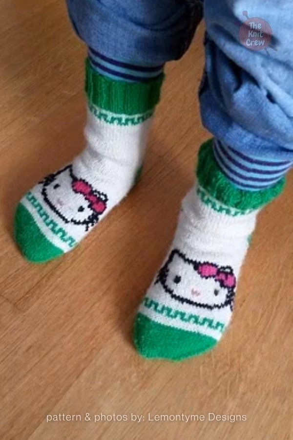 A pair of white and green knitted socks being worn, showcased in a collection from The Knit Crew. The socks feature a bright green cuff, heel, and toe, with a decorative geometric green border. On the top of the foot, there is a detailed knit design of Hello Kitty&rsquo;s face with her signature pink bow. The image is part of a curated pattern roundup for kitty-themed gifts.