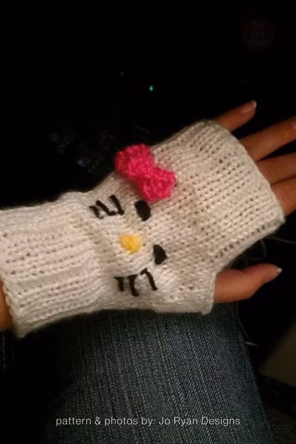A hand wearing a white knitted fingerless glove, featured in a design roundup from The Knit Crew. The glove is made with a simple knit stitch and ribbed cuff, adorned with an embroidered Hello Kitty face. The face includes black oval eyes, black whiskers, a yellow nose, and a prominent 3D hot pink crochet bow attached near the top.