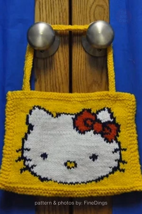 A bright yellow knitted tote bag hanging from door handles, part of a pattern collection from The Knit Crew. The center of the bag features a large intarsia or duplicate stitch design of Hello Kitty&rsquo;s face in white with black outlines. She is wearing a classic red bow with white polka dots, and the bag is finished with two sturdy yellow knitted handles.