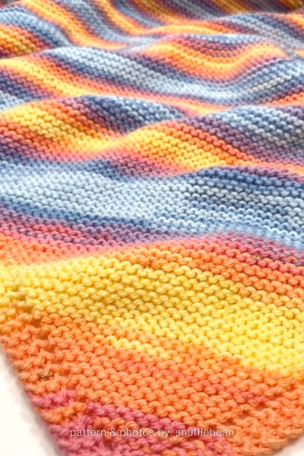 A close-up photo of a softly folded pastel rainbow striped knitted blanket, showing bands of peach, yellow, light blue, and purple yarn in a garter stitch pattern, featured in a knitting roundup from https://www.theknitcrew.com/.