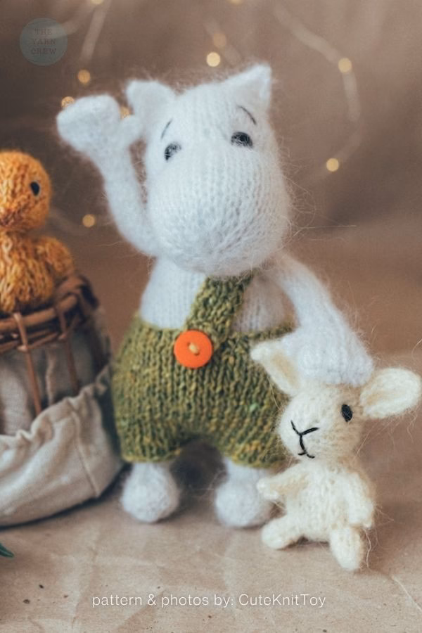 A white knitted hippo toy standing upright with one arm raised in a wave, wearing green knitted overalls with a single orange button. Beside it stands a tiny white knitted lamb toy. In the blurred background, there is a small orange knitted bird in a basket and warm fairy lights. Text at the bottom reads "pattern & photos by: CuteKnitToy". This is part of a knitting roundup from The Yarn Crew.