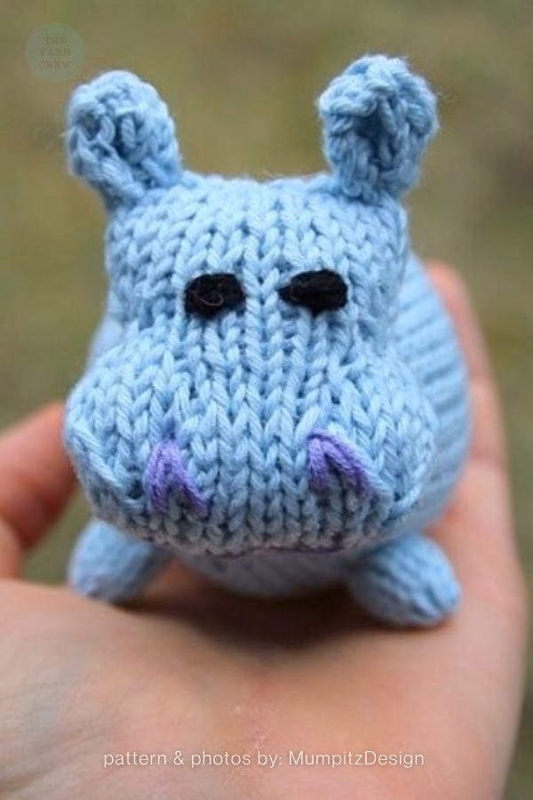 A close-up of a small, light blue knitted hippo toy resting in the palm of a hand. The hippo features black stitched eyes and purple stitched nostrils. Text at the bottom reads "pattern & photos by: MumpitzDesign". This is part of a knitting roundup from The Yarn Crew.