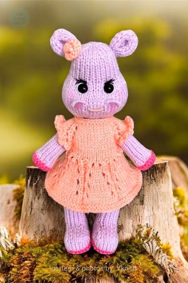 A purple knitted hippo toy standing upright on a wooden stump outdoors. It is wearing a peach-colored knitted dress with ruffled sleeves and has a small flower accessory on its ear. Text at the bottom reads "pattern & photos by: VknitB". This is part of a knitting roundup from The Yarn Crew.