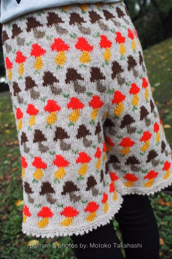 A close-up view of light beige knitted adult shorts featuring an all-over fair isle pattern of red, yellow, and brown mushrooms. Curated by Ariana of TheKnitCrew.