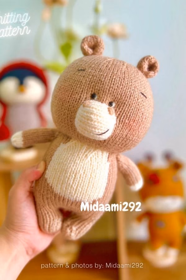 A hand holding a light brown knitted hippo toy with a cream-colored belly and snout. The toy has small stitched eyes, a smiling mouth, and small round ears. Text overlay reads "Midaami292" and "pattern & photos by: Midaami292" at the bottom. This is part of a crochet roundup from https://www.theknitcrew.com/.