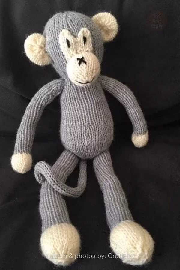 A slender, grey knitted monkey plush toy with cream-colored hands, feet, ears, and snout, sitting against a dark background. Curated by Ariana of TheKnitCrew.