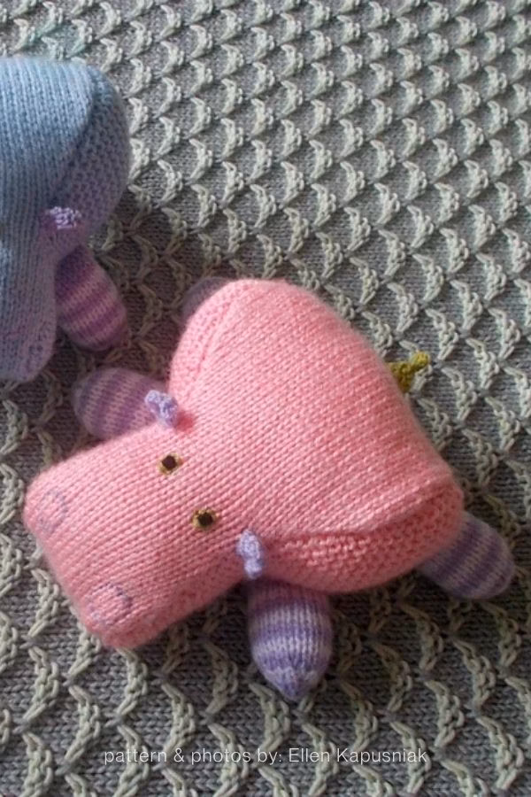 A small, flat pink knitted hippo toy with purple and white striped legs and tiny purple ears, resting on a grey textured knitted blanket. A partial view of a similar blue hippo is visible in the background. Text reads "pattern & photos by: Ellen Kapusniak". This is part of a crochet roundup from https://www.theknitcrew.com/.