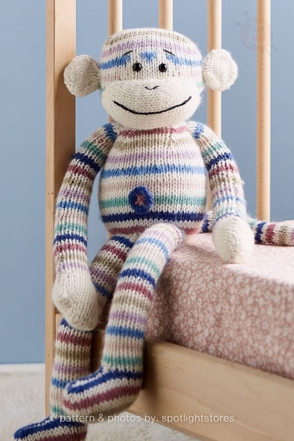 A colorful, multi-striped knitted monkey plush with a cream face, sitting on a pink patterned mattress next to a wooden crib. Curated by Ariana of TheKnitCrew.