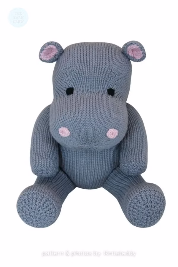 A grey knitted hippo toy sitting upright against a plain white background. It features pink inner ears, pink stitched blush spots on its cheeks, and black stitched eyes. Text at the bottom reads "pattern & photos by: Knitateddy". This is part of a crochet roundup from https://www.theknitcrew.com/.