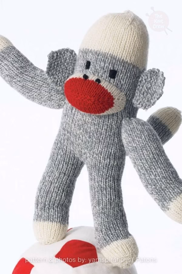 A classic grey and white knitted sock monkey plush with a bright red mouth, standing on a red and white patterned surface. Curated by Ariana of TheKnitCrew.