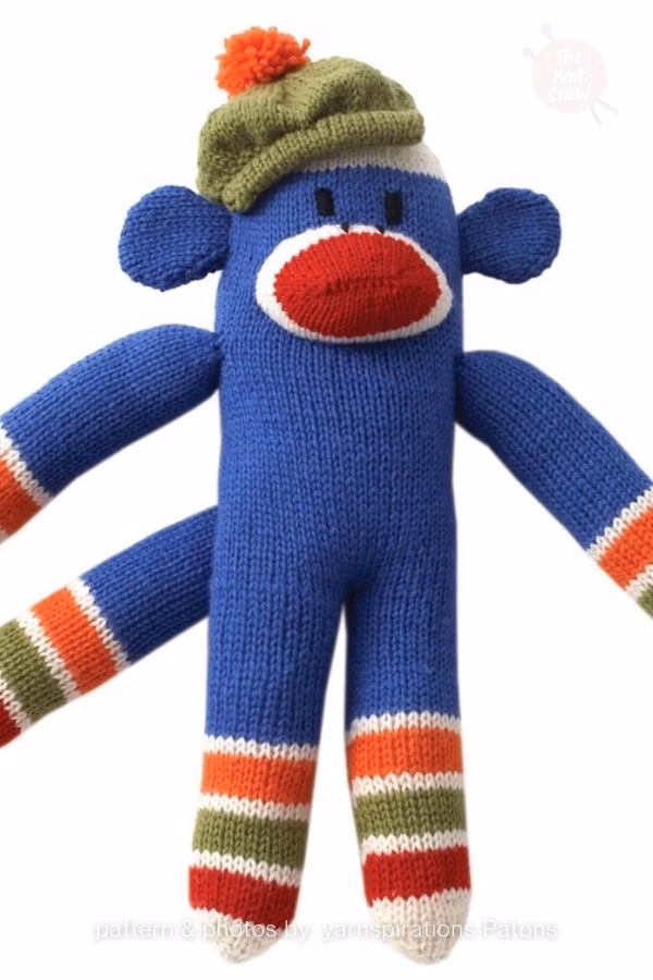 A vibrant blue knitted sock monkey plush wearing a green beanie with an orange pom-pom, featuring striped arms and legs. Curated by Ariana of TheKnitCrew.