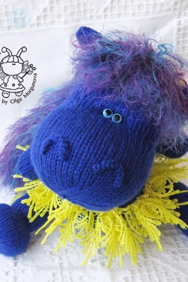 A bright blue knitted hippo toy with a fluffy purple and blue yarn mane, bright blue safety eyes, and a neon yellow lace collar. It is resting on a white patterned cloth. A logo and text read "pattern & photos by: Morgunova Olga". This is part of a crochet roundup from https://www.theknitcrew.com/.