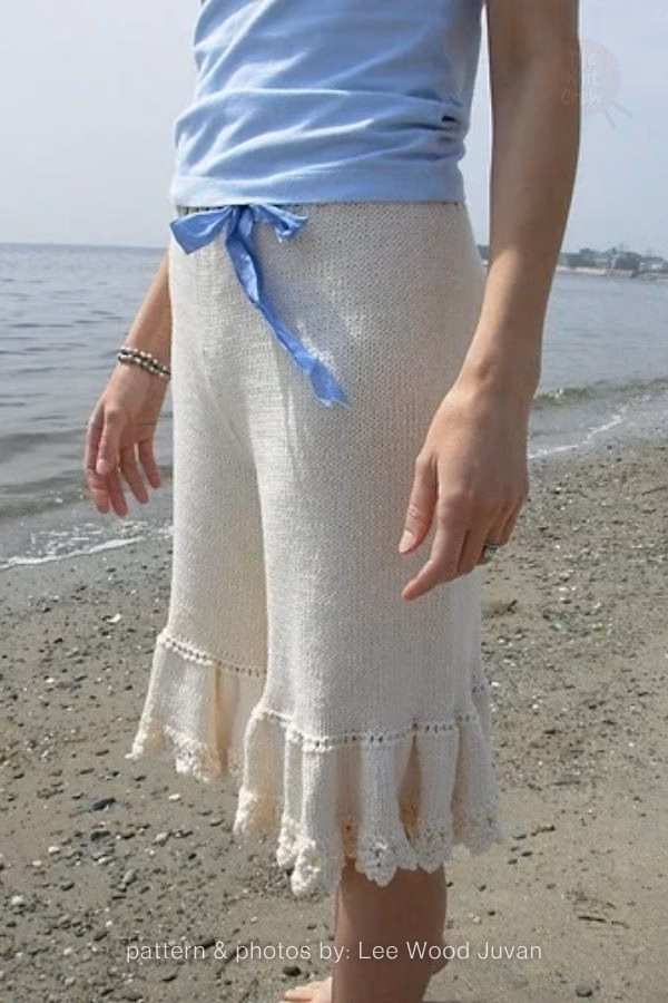 A person on a beach wearing long, flowing off-white knitted adult shorts with a large ruffled hem and a blue ribbon waist tie. Curated by Ariana of TheKnitCrew.