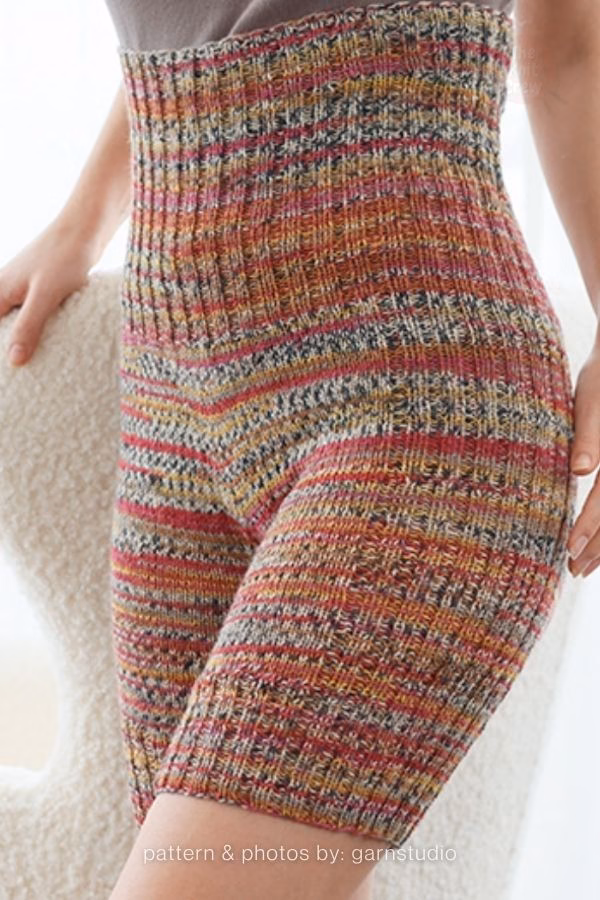 A close-up view of high-waisted knitted adult shorts featuring a multi-colored variegated yarn pattern with red, orange, yellow, and grey stripes. Curated by Ariana of TheKnitCrew.