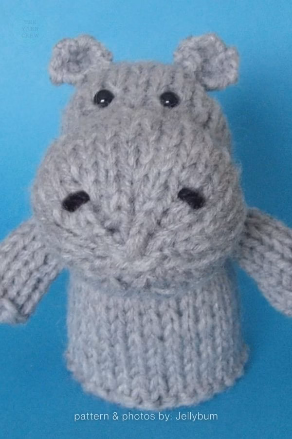 A close-up of a grey knitted hippo toy with small ears, black stitched eyes, and black stitched nostrils, set against a solid blue background. Text at the bottom reads "pattern & photos by: Jellybum". This is part of a knitting roundup from The Yarn Crew.
