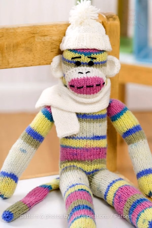 A knitted monkey plush with colorful pink, blue, and yellow stripes wearing a white winter beanie and a matching white scarf. Curated by Ariana of TheKnitCrew.