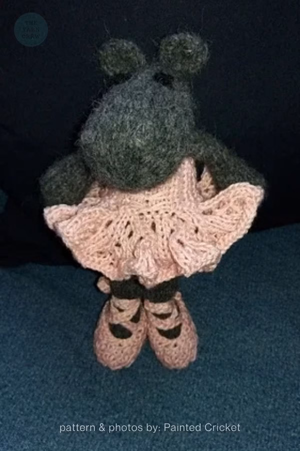 A dark grey knitted hippo toy standing upright, wearing a frilly light pink knitted dress and matching pink ballet-style shoes with dark straps. It is posed against a dark background. Text at the bottom reads "pattern & photos by: Painted Cricket". This is part of a knitting roundup from The Yarn Crew.
