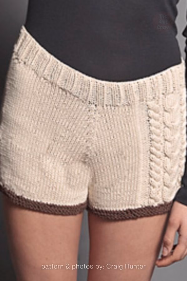 A close-up view of textured cream-colored knitted adult shorts featuring a braided cable stitch detail down the side and dark brown ribbed edging along the hem. Curated by Ariana of TheKnitCrew.