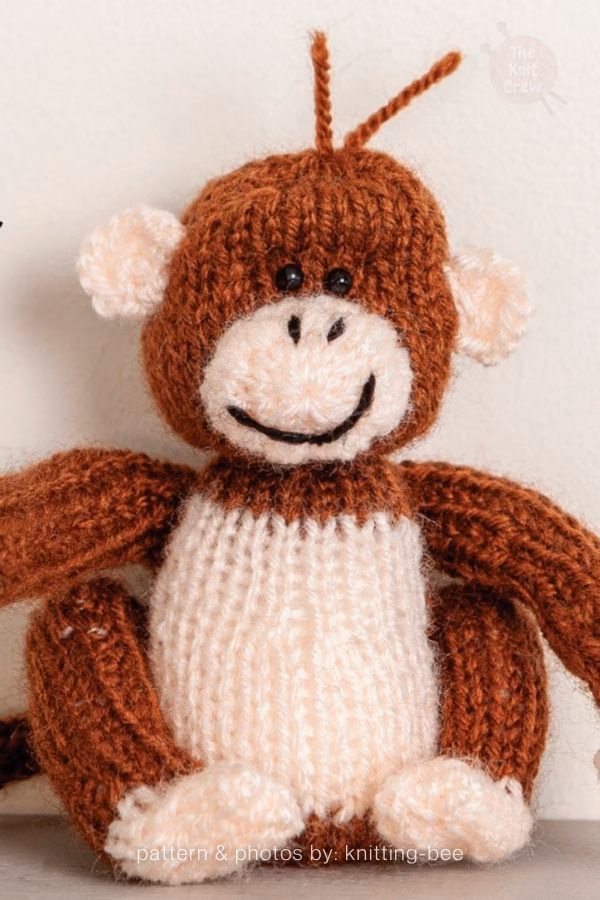 A fluffy brown knitted monkey plush with a cream belly, face, and ears, featuring a small yarn topknot strand on its head. Curated by Ariana of TheKnitCrew.