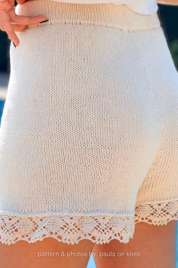 A back view of simple light cream knitted adult shorts featuring a delicate, intricate lace trim along the leg hems. Curated by Ariana of TheKnitCrew.
