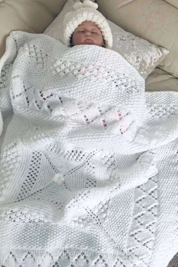 A sleeping baby wearing a knit hat tucked under a textured white knitted blanket with a mix of geometric openwork and eyelet stitch patterns. Text at the bottom reads: "pattern & photos by: HoneyBeeBears". Curated by Ariana of TheKnitCrew.