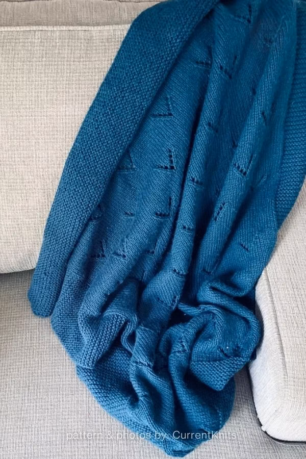 A rich teal or dark blue knitted blanket draped loosely over the arm of a beige or grey sofa, featuring small, scattered pointelle eyelet accents. Text at the bottom reads: "pattern & photos by: CurrentKnits". Curated by Ariana of TheKnitCrew.