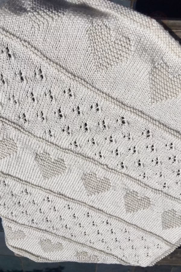 A close-up view of an off-white knitted blanket featuring alternating horizontal bands of eyelet pointelle lace and textured knit hearts. Text at the bottom reads: "pattern & photos by: KnitSewMake". Curated by Ariana of TheKnitCrew.