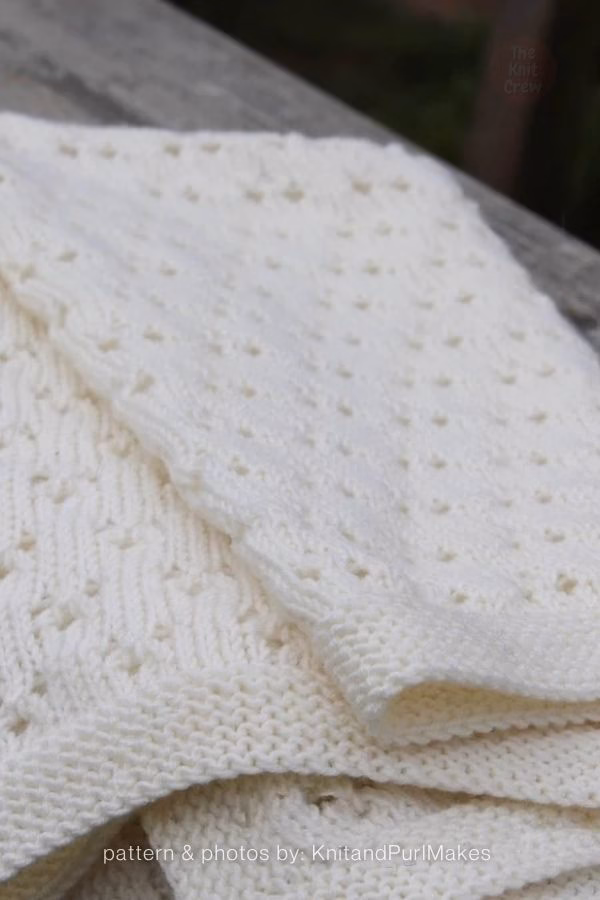A detailed close-up of a folded, cream-white knitted blanket showcasing intricate zigzag textured stitch patterns and subtle lace eyelets. Text at the bottom reads: "pattern & photos by: KnitandPurlMakes". Curated by Ariana of TheKnitCrew.