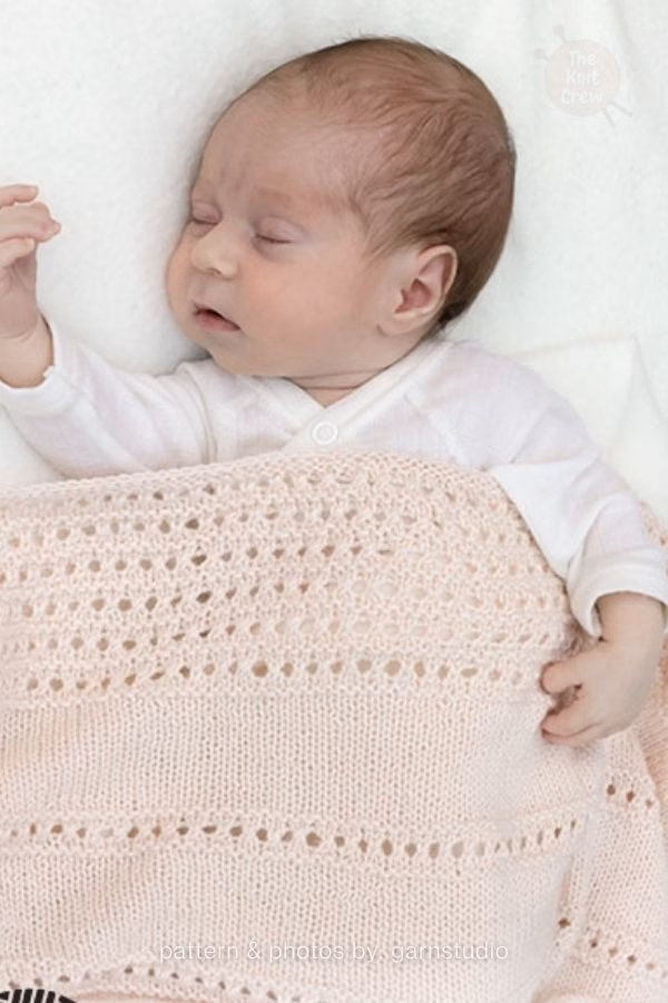 A sleeping baby tucked under a soft, light pink knitted blanket featuring rows of delicate horizontal lace eyelet bands. Text at the bottom reads: "pattern & photos by: garnstudio". Curated by Ariana of TheKnitCrew.