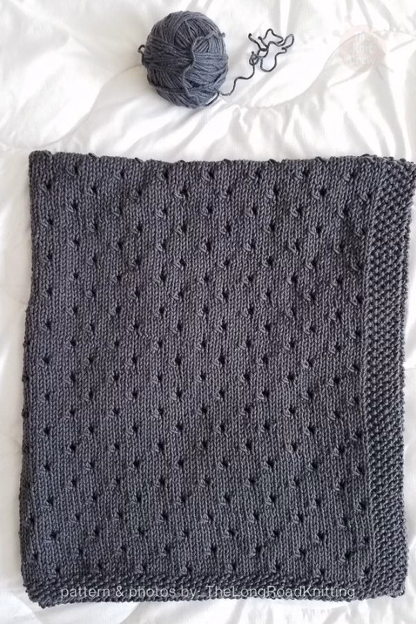 A dark grey knitted blanket laid flat on a white surface, displaying a dense pattern of small eyelet details and a seed stitch border. A matching ball of grey yarn sits at the top. Text at the bottom reads: "pattern & photos by: TheLongRoadKnitting". Curated by Ariana of TheKnitCrew.