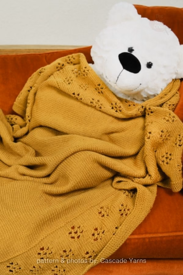 A mustard-yellow knitted blanket draped over an orange chair next to a white plush teddy bear. The blanket features delicate leaf-like lace border accents. Text at the bottom reads: "pattern & photos by: Cascade Yarns". Curated by Ariana of TheKnitCrew.