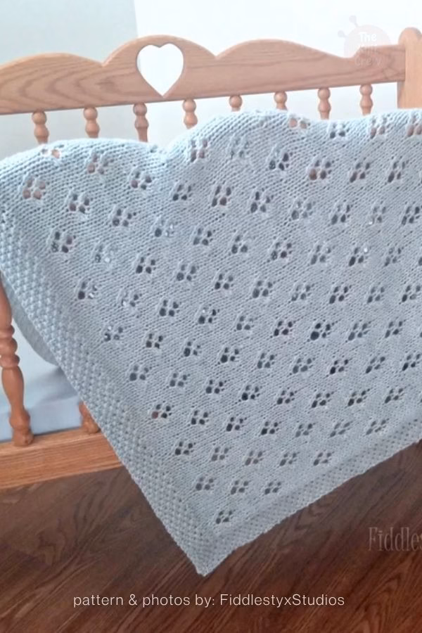 A light blue or grey baby blanket with an all-over repeating diamond eyelet pattern draped over a wooden crib. Text at the bottom reads: "pattern & photos by: FiddlestyxStudios". Curated by Ariana of TheKnitCrew.