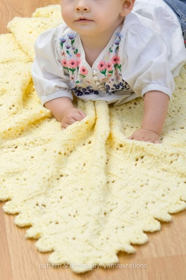 A baby in an embroidered floral top lying on a soft, pale yellow knitted blanket featuring wavy lace panels and a scalloped edge. Text at the bottom reads: "pattern & photos by: yarnspirations". Curated by Ariana of TheKnitCrew.