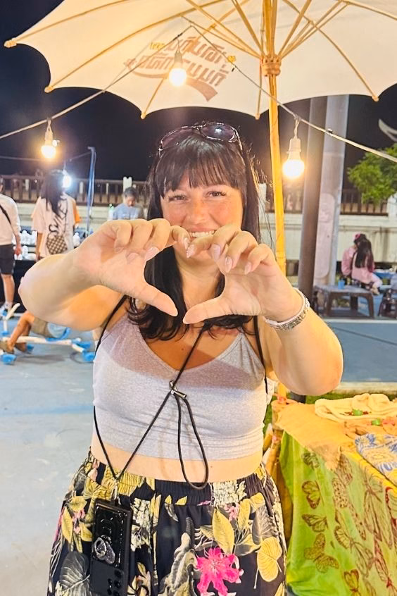 Ariana, pattern curator @TheKnitCrew and crochet designer, smiling and making a heart shape with her hands at an outdoor market under a lit umbrella.