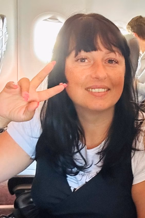 Ariana, pattern curator @TheKnitCrew and crochet designer, smiling and making a peace sign with her hand while sitting by a window inside an airplane cabin.