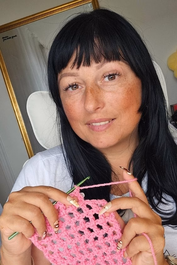 Ariana @TheKnitCrew, designer and pattern curator, smiling while crocheting a pink project with a green crochet hook in her seaside studio.