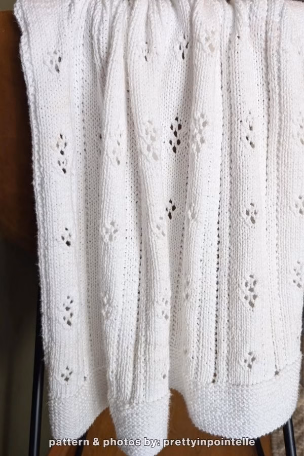 A white knitted blanket in pointelle pattern draped over the chair. Text at the bottom reads: "pattern & photos by: prettyinpointelle". Curated by Ariana of TheKnitCrew.